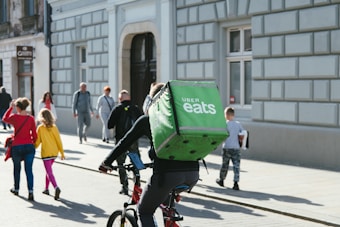 A bustling urban street scene with a cyclist carrying an Uber Eats delivery bag riding past pedestrians walking along the sidewalk. The street features a historical building facade with large windows and entranceways, while the people are dressed in casual attire, suggesting a typical day in the city.