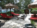 A serene outdoor setting features several red-cushioned lounge chairs beneath straw umbrellas. These are arranged along a stone pathway surrounded by lush green trees and plants. In the background, there's a glimpse of a blue ocean, creating a relaxing and tropical atmosphere.