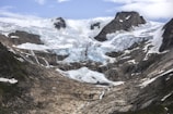 A majestic mountainous landscape features a massive glacier flowing down between rugged peaks. The ice is predominantly white with patches of blue, surrounded by dark, jagged rock formations. Sparse patches of greenery are visible in places where the snow or ice has receded.