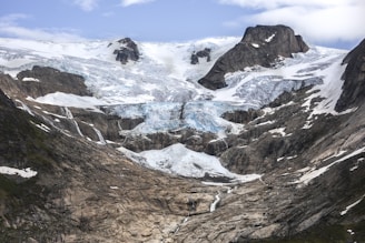 A majestic mountainous landscape features a massive glacier flowing down between rugged peaks. The ice is predominantly white with patches of blue, surrounded by dark, jagged rock formations. Sparse patches of greenery are visible in places where the snow or ice has receded.