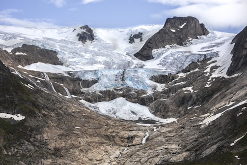 A majestic mountainous landscape features a massive glacier flowing down between rugged peaks. The ice is predominantly white with patches of blue, surrounded by dark, jagged rock formations. Sparse patches of greenery are visible in places where the snow or ice has receded.