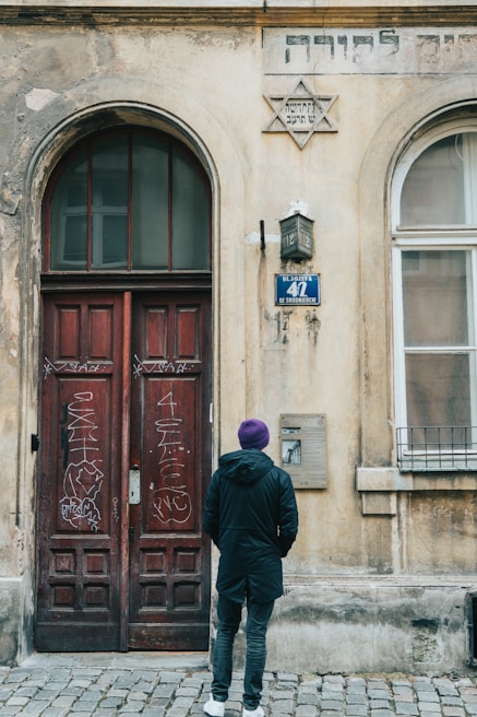 A person wearing a purple hat and dark coat stands in front of an old building with a large, weathered wooden door adorned with graffiti. Above the door, there is a Star of David and inscriptions in Hebrew. The building's facade appears aged with cracks and faded paint. A street sign with the number 41 and a plaque are also visible.