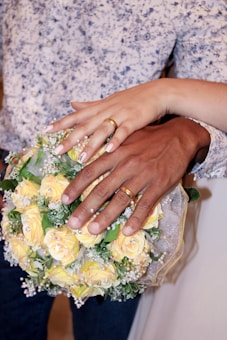 Two hands, each wearing a gold wedding ring, rest on a bouquet of yellow roses. One hand is lighter-skinned, and the other is darker-skinned. The bouquet is adorned with small white flowers and greenery. Part of a shirt with a blue floral pattern and a white garment are visible in the background.