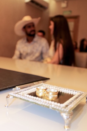 A blurred couple sits in the background, with a decorative tray holding two gold rings prominently displayed in the foreground. The setting appears to be a formal or ceremonial occasion, suggested by the elegant presentation of the rings.