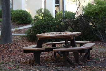 A wooden picnic table is surrounded by fallen autumn leaves in a park setting. The background features green bushes and a tree trunk, with a staircase and light-colored buildings visible in the distance.