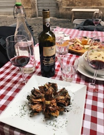 A rustic outdoor dining setup featuring a red and white checkered tablecloth. A centerpiece of fried artichoke is elegantly placed on a white square plate, garnished with herbs. Nearby, a bottle of Chianti Classico and a glass of red wine are complemented by two clear water glasses. A platter of assorted appetizers, including cured meats and cheese, is visible in the background.