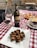 A rustic outdoor dining setup featuring a red and white checkered tablecloth. A centerpiece of fried artichoke is elegantly placed on a white square plate, garnished with herbs. Nearby, a bottle of Chianti Classico and a glass of red wine are complemented by two clear water glasses. A platter of assorted appetizers, including cured meats and cheese, is visible in the background.