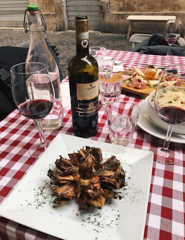 A rustic outdoor dining setup featuring a red and white checkered tablecloth. A centerpiece of fried artichoke is elegantly placed on a white square plate, garnished with herbs. Nearby, a bottle of Chianti Classico and a glass of red wine are complemented by two clear water glasses. A platter of assorted appetizers, including cured meats and cheese, is visible in the background.