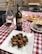 A rustic outdoor dining setup featuring a red and white checkered tablecloth. A centerpiece of fried artichoke is elegantly placed on a white square plate, garnished with herbs. Nearby, a bottle of Chianti Classico and a glass of red wine are complemented by two clear water glasses. A platter of assorted appetizers, including cured meats and cheese, is visible in the background.