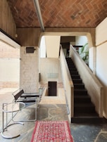 A modern interior space featuring a staircase with a minimalist design. The area includes exposed brickwork, concrete walls, and a sloped ceiling. There's a chair with a sleek metal frame and a round glass-topped table. A decorative red and patterned rug adds a warm touch to the stone floor.