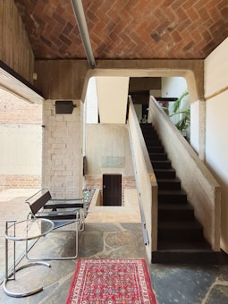 A modern interior space featuring a staircase with a minimalist design. The area includes exposed brickwork, concrete walls, and a sloped ceiling. There's a chair with a sleek metal frame and a round glass-topped table. A decorative red and patterned rug adds a warm touch to the stone floor.