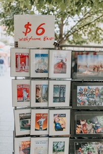 A display stand holds various art prints and posters in frames. Each print features colorful and diverse artwork, including cityscapes and famous magazine covers. A sign on top reads '$6 with frame 8x10'. The background shows leafy trees.