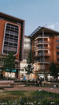 A modern building with large windows and multiple floors stands against a clear sky. The structure features a combination of glass and brick elements, with a geometric design and a rooftop solar panel. In front of the building, well-maintained greenery and a small sculpture add to the urban landscape, and a few people can be seen near the entrance.