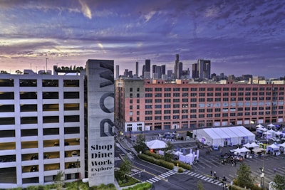 An urban scene with a large parking structure and modern city buildings. The foreground features a sign with 'ROW' and 'Shop Dine Work Explore' written on it. Tents and booths are set up in an open area, suggesting a market or event. The skyline in the background consists of tall skyscrapers under a dramatic evening sky.
