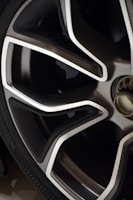 Close-up view of a modern car wheel rim with a sleek design. The rim features a combination of metallic silver and dark gray colors with sharp, angular lines. The tire is also partially visible in the image.