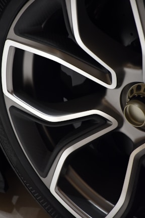Close-up view of a modern car wheel rim with a sleek design. The rim features a combination of metallic silver and dark gray colors with sharp, angular lines. The tire is also partially visible in the image.