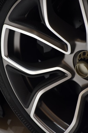 Close-up view of a modern car wheel rim with a sleek design. The rim features a combination of metallic silver and dark gray colors with sharp, angular lines. The tire is also partially visible in the image.