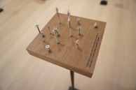 A wooden block on a stand features an arrangement of screws of varying heights. Small white figurines are perched atop several screws, creating an impression of staggered levels. The setup casts shadows on the wood surface, and there is an engraved text on the block. The background is a smooth, neutral tone, lending focus to the structure.