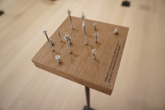 A wooden block on a stand features an arrangement of screws of varying heights. Small white figurines are perched atop several screws, creating an impression of staggered levels. The setup casts shadows on the wood surface, and there is an engraved text on the block. The background is a smooth, neutral tone, lending focus to the structure.