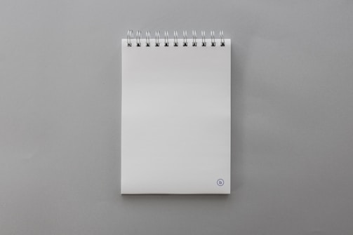 A blank, white spiral-bound notepad placed on a smooth gray surface. The notepad features a small, circular logo at the bottom right corner on the front page.