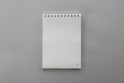 A blank, white spiral-bound notepad placed on a smooth gray surface. The notepad features a small, circular logo at the bottom right corner on the front page.