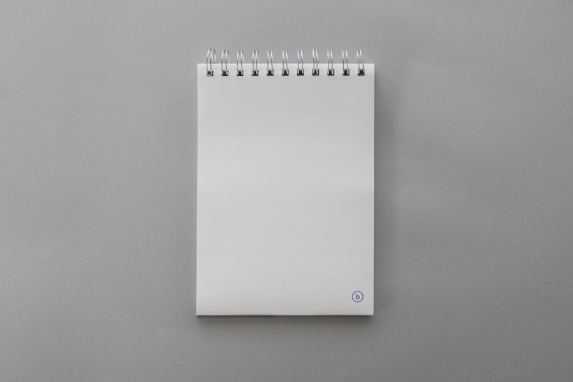 A blank, white spiral-bound notepad placed on a smooth gray surface. The notepad features a small, circular logo at the bottom right corner on the front page.