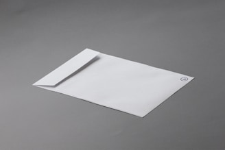 A white envelope with a minimalistic design lies on a smooth gray surface. The envelope features a simple logo or emblem in the top corner.