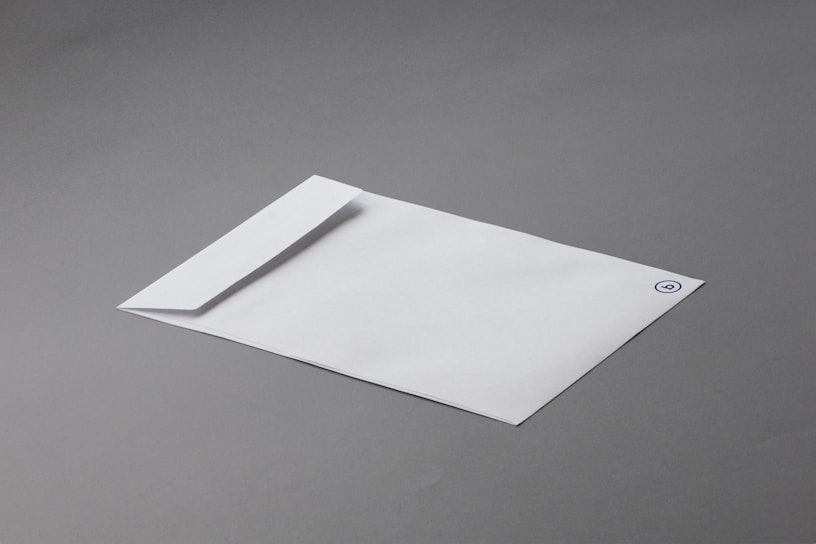 A white envelope with a minimalistic design lies on a smooth gray surface. The envelope features a simple logo or emblem in the top corner.
