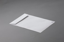 A white envelope with a minimalistic design lies on a smooth gray surface. The envelope features a simple logo or emblem in the top corner.