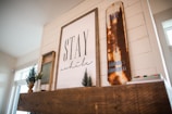 A cozy interior setting featuring a wooden mantel adorned with rustic decor. Centered on the mantel is a large framed artwork with the text 'Stay a while.' To the right, a vintage, distressed sign reads 'Canada's Favourite,' with visible rust giving it a nostalgic feel. Small potted plants add a touch of greenery, enhancing the warm and inviting atmosphere.