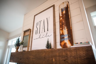 A cozy interior setting featuring a wooden mantel adorned with rustic decor. Centered on the mantel is a large framed artwork with the text 'Stay a while.' To the right, a vintage, distressed sign reads 'Canada's Favourite,' with visible rust giving it a nostalgic feel. Small potted plants add a touch of greenery, enhancing the warm and inviting atmosphere.