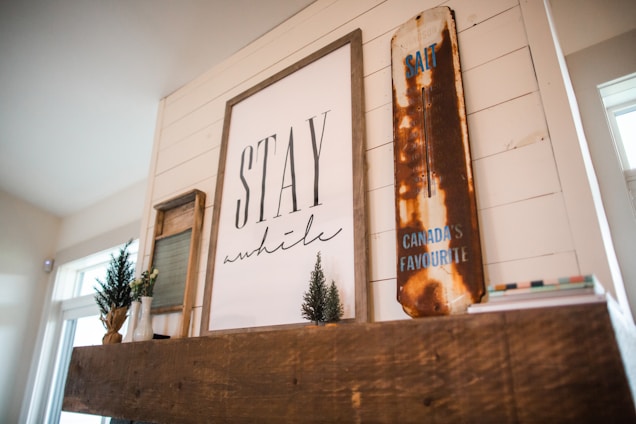 A cozy interior setting featuring a wooden mantel adorned with rustic decor. Centered on the mantel is a large framed artwork with the text 'Stay a while.' To the right, a vintage, distressed sign reads 'Canada's Favourite,' with visible rust giving it a nostalgic feel. Small potted plants add a touch of greenery, enhancing the warm and inviting atmosphere.