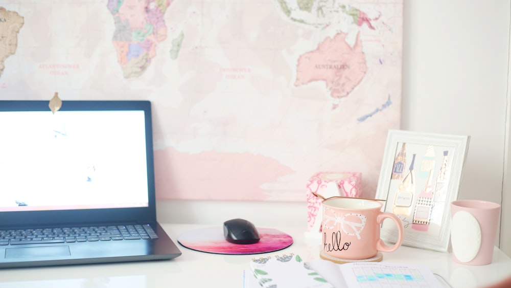 desk with turned-on laptop, mouse, mousepad and cup of coffee