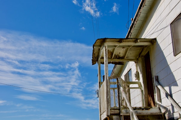 A weathered and aged wooden structure forms the porch of an old white house, showing signs of decay. The porch is positioned against a backdrop of a bright blue sky with scattered white clouds. The wooden materials are visibly deteriorating, and electrical wires run above the building.
