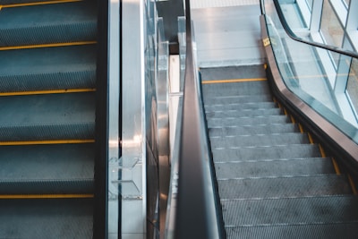 A pair of adjacent escalators, one going up and the other going down, in a modern indoor environment. The escalator steps are grey with yellow safety edges, and there are reflective metal and glass elements on the sides. Natural light illuminates the scene through large windows.