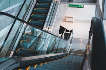 An empty escalator with a clean and modern design descends to the lower level of a building. The area is well-lit with natural and artificial lighting, featuring a green sign indicating directions to a mall link. The walls are smooth and light-colored, complementing the reflective glass sides of the escalator.