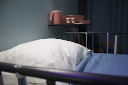 A hospital bed with a white pillow and blue blanket is in the foreground. On the wall shelf, there are pink containers including a pitcher and two bottles. The setting appears to be a medical facility with a calm atmosphere.