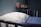 A hospital bed with a white pillow and blue blanket is in the foreground. On the wall shelf, there are pink containers including a pitcher and two bottles. The setting appears to be a medical facility with a calm atmosphere.