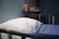 A hospital bed with a white pillow and blue blanket is in the foreground. On the wall shelf, there are pink containers including a pitcher and two bottles. The setting appears to be a medical facility with a calm atmosphere.