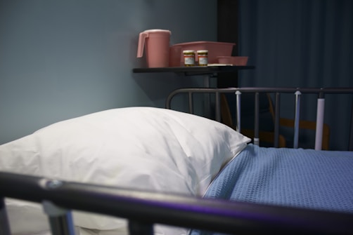 A hospital bed with a white pillow and blue blanket is in the foreground. On the wall shelf, there are pink containers including a pitcher and two bottles. The setting appears to be a medical facility with a calm atmosphere.