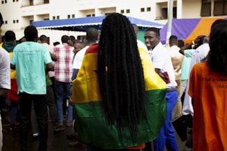 A crowd of people is gathered outdoors, with a noticeable individual wrapped in a vibrant flag that features yellow, green, and red stripes with a black star. The scene includes a mix of people wearing plain and patterned shirts, some of whom are facing away from the camera. The backdrop includes a building and a tent with blue and orange colors.