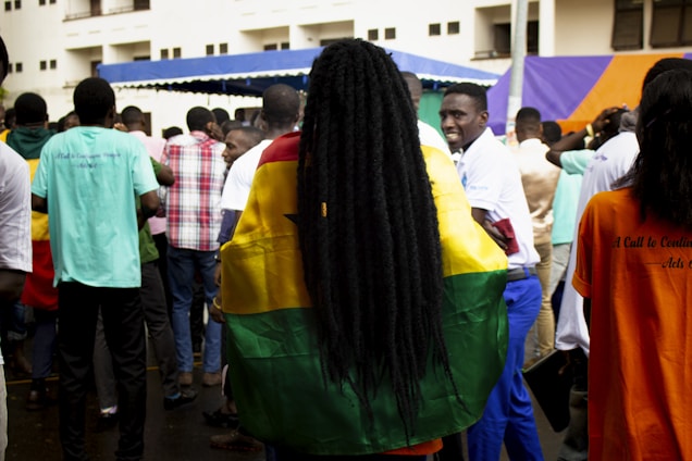 A crowd of people is gathered outdoors, with a noticeable individual wrapped in a vibrant flag that features yellow, green, and red stripes with a black star. The scene includes a mix of people wearing plain and patterned shirts, some of whom are facing away from the camera. The backdrop includes a building and a tent with blue and orange colors.