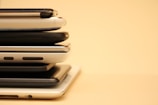 A stack of smartphones piled on top of one another, with a variety of cases and designs, set against a beige background. The phones vary in colors and styles, showing a progression of technological design.