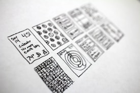 A series of hand-drawn sketches on a piece of paper, featuring various rectangles filled with doodles. The sketches include a calendar with written text indicating a specific date and time, abstract diagrams, and grid patterns with numbers or symbols.