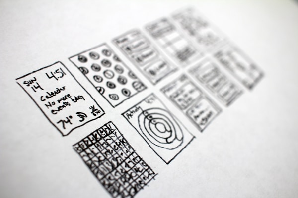 A series of hand-drawn sketches on a piece of paper, featuring various rectangles filled with doodles. The sketches include a calendar with written text indicating a specific date and time, abstract diagrams, and grid patterns with numbers or symbols.