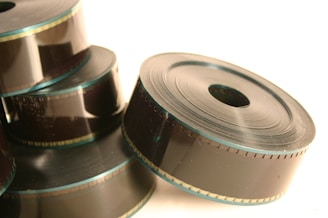 Several large rolls of film are stacked together, with a glossy texture reflecting light. The film appears to be wound tightly, with visible edges and perforations along one side. The overall setup is simple, capturing the detail of the film’s design.