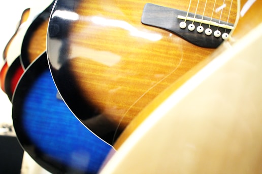 A close-up view of the bodies of several acoustic guitars with visible strings and a glossy finish. The guitars display different colors, including sunburst, blue, and natural wood tones.