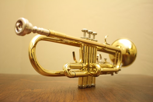 A shiny brass trumpet rests on a wooden surface, showcasing its intricate details and glossy finish. The background is a soft, neutral color that highlights the instrument’s metallic surface.