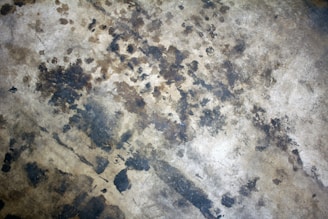A worn and weathered concrete surface featuring various dark stains and discolorations throughout. The texture is rough and uneven, with patches of darker brown and black scattered across.