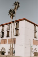 A white building with a Spanish architectural style features red-tiled roofs and multiple balconies with iron railings. Tall palm trees stand in front of the building, and there is minimal shadowing on the walls. The corner of the building shows street signs and is labeled 'The Village' and numbered '1418'.